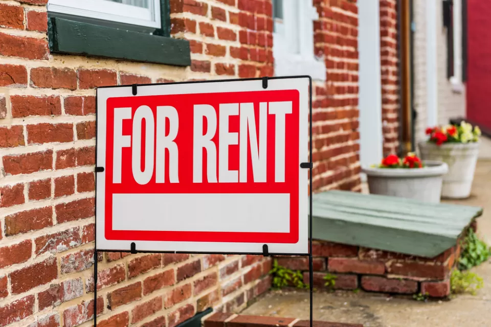 Red and white For Rent sign in front of brick building with flowerpots in background
